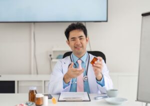 Médico sorridente segura frasco de medicamento, sentado à mesa com prontuário e objetos médicos, simbolizando posicionamento digital com foco educativo.